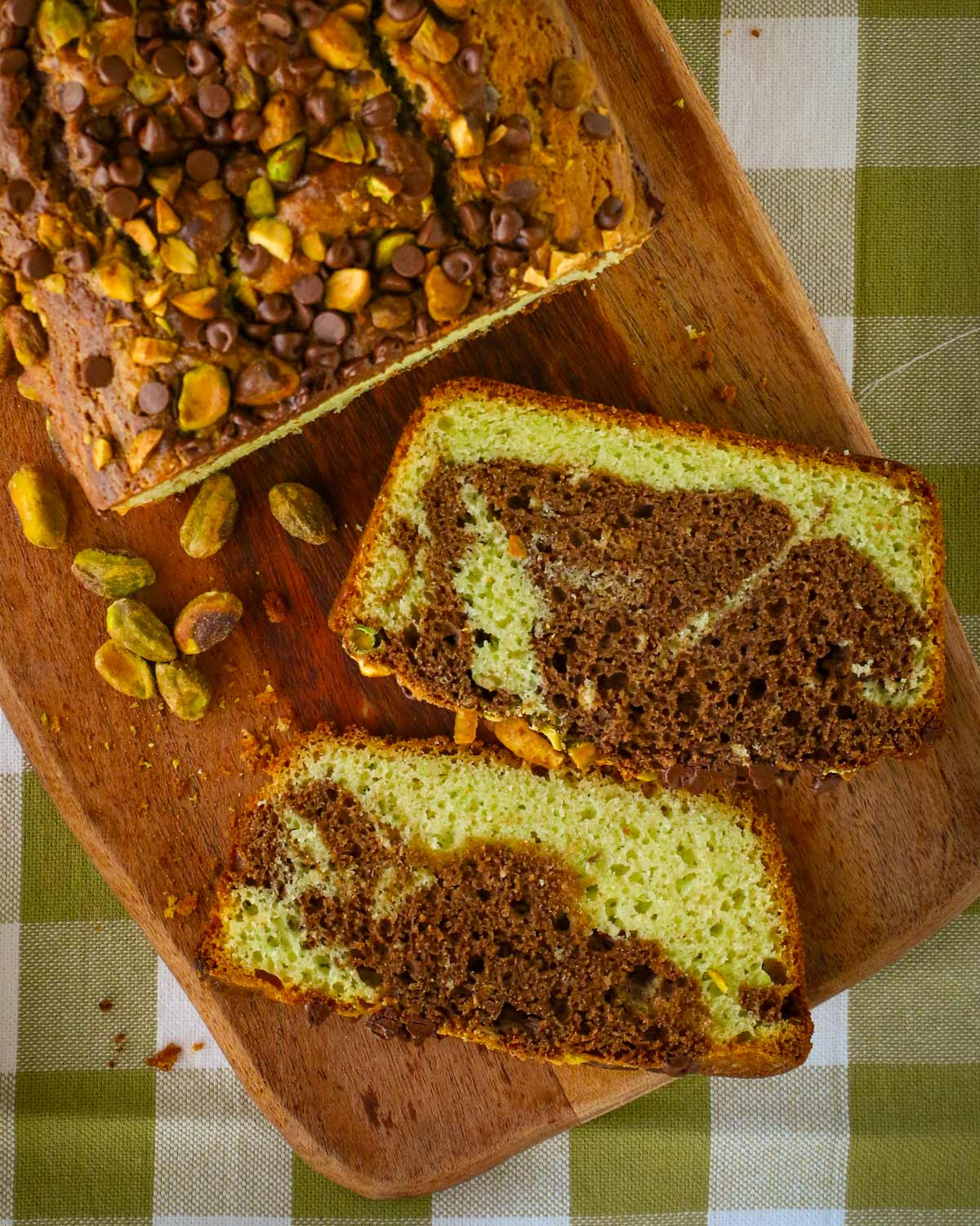 slices of chocolate pistachio loaf on a wooden board on a green checked cloth.