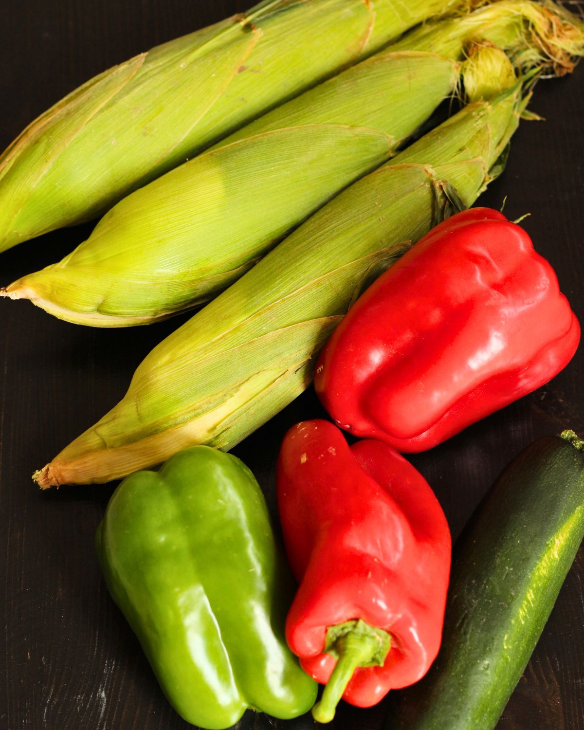 ears of corn, bell peppers, and a zucchini on a black table top.